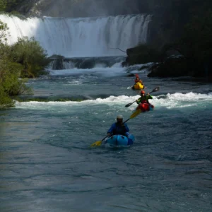 Three people in packrafts, paddling down Zrmanja river with the waterfall in the background