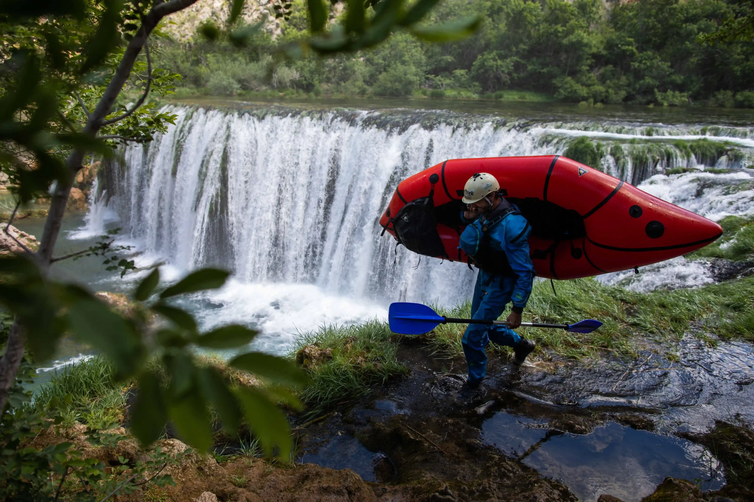 A person walking above a big waterfall on Zrmanja river while carrying a paddle and a packraft.
