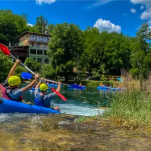 2 osobe u kajaku idu niz slap na Mrežnici sa restoranom "Zeleni Kut" u pozadini